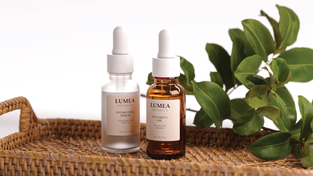 Botanical Skincare Still Life — wide view of two dropper bottles on a woven tray with green leaves